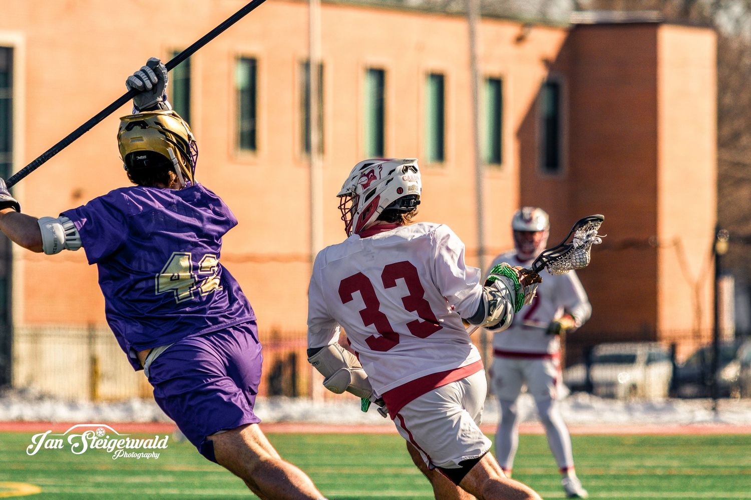 Two lacrosse players battle for possession during an intense game at an outdoor field with brick buildings in background.