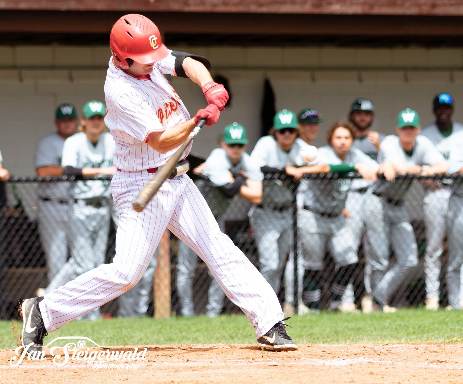 Baseball player in white pinstriped uniform swings bat at home plate while opposing team watches from dugout.