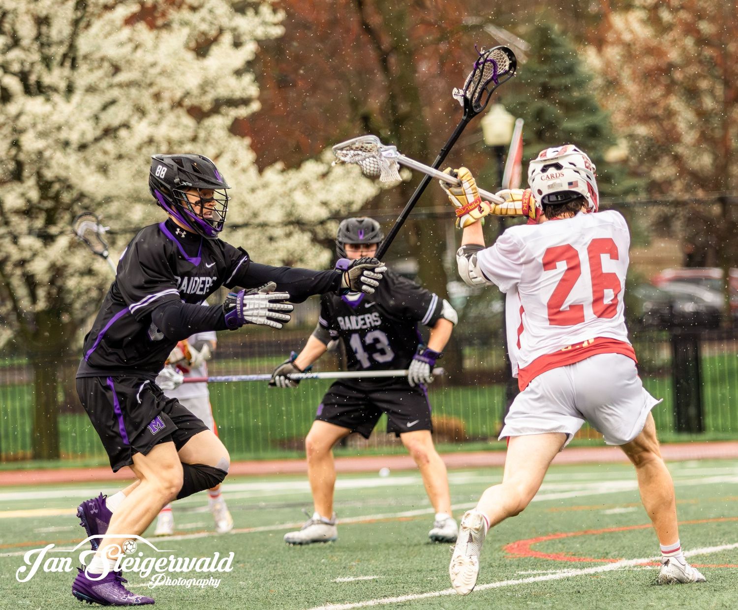 Lacrosse players in black and white uniforms compete intensely during a match with spring blossoms in background.