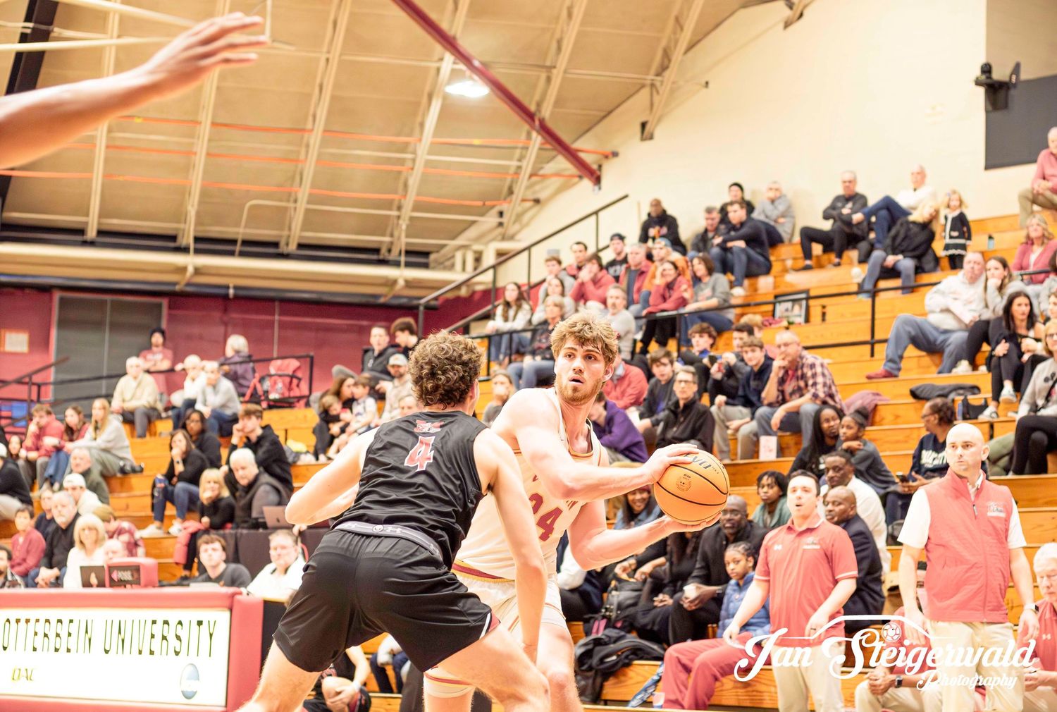 Basketball players leap high to block a shot during an intense college game with spectators watching from the bleachers.