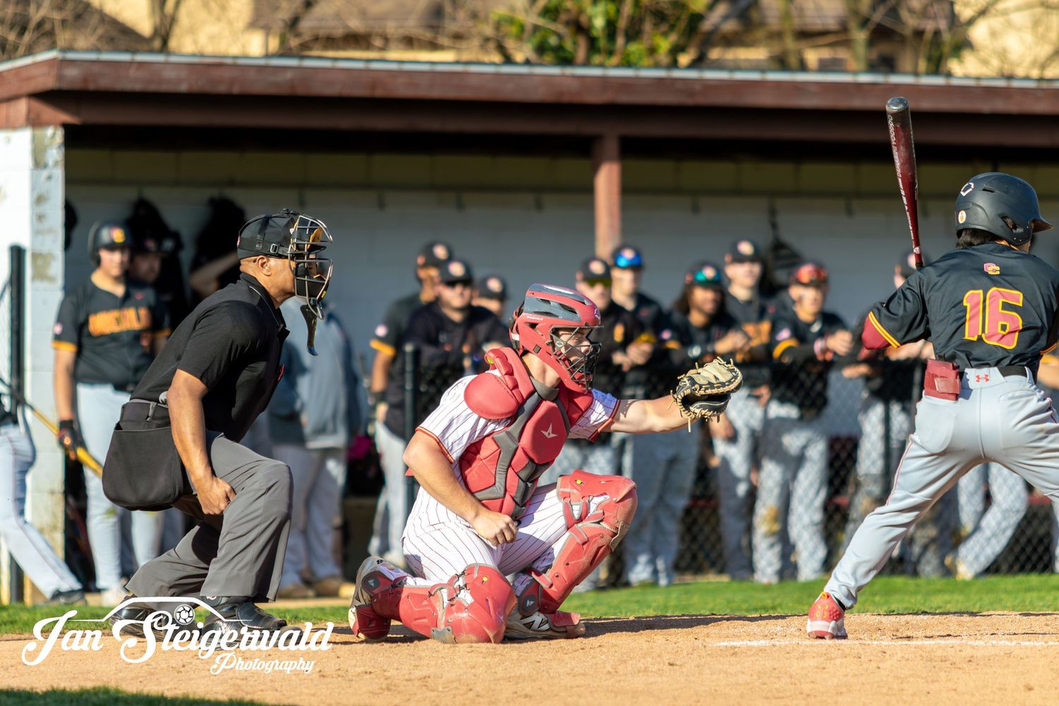 Baseball batter at plate with catcher crouched behind during game as opposing team watches from dugout.