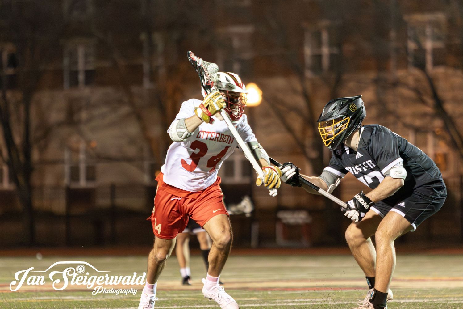 Two lacrosse players battle for possession during intense night game with player in red attacking against black defender.