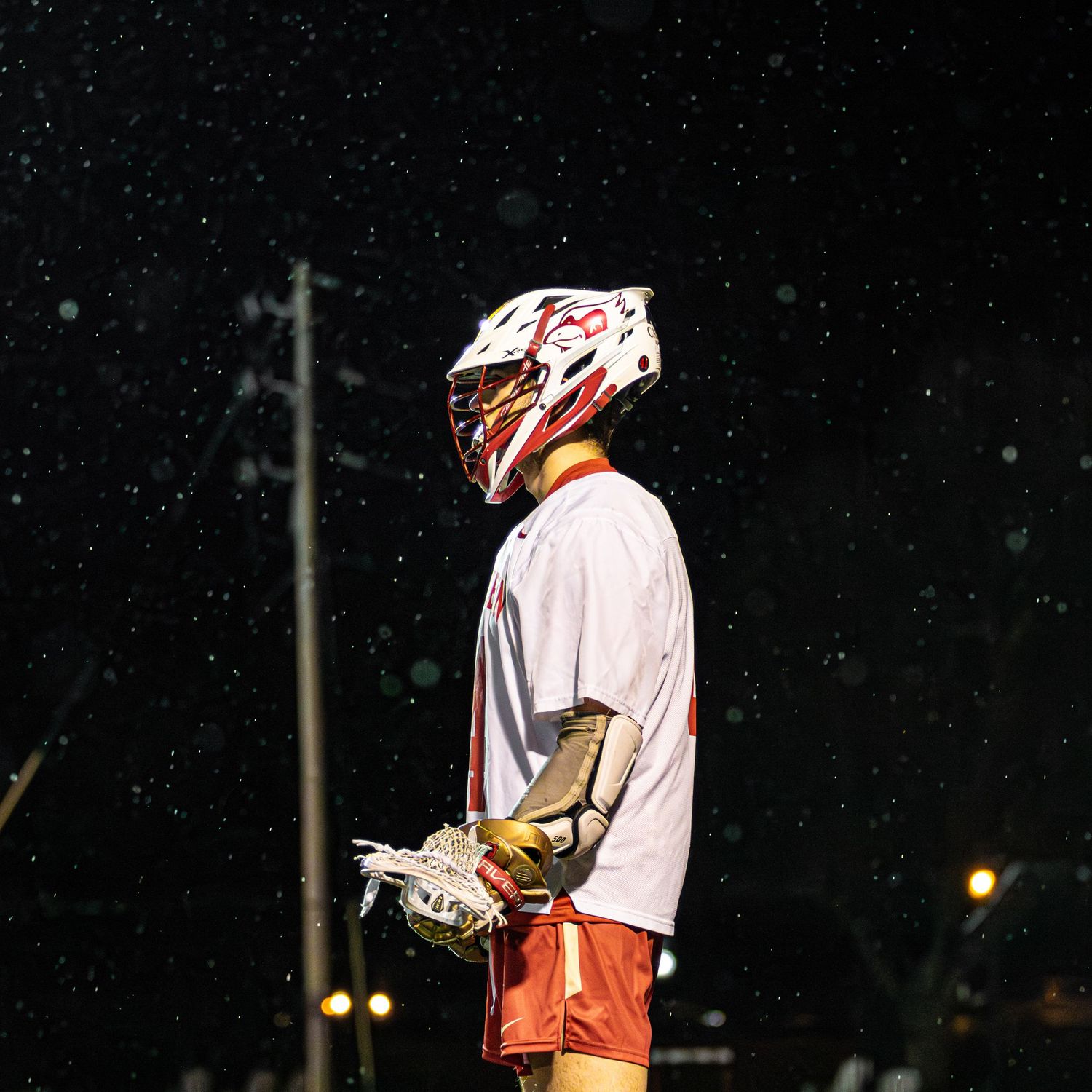 Lacrosse player in white uniform and red helmet standing under night lights.