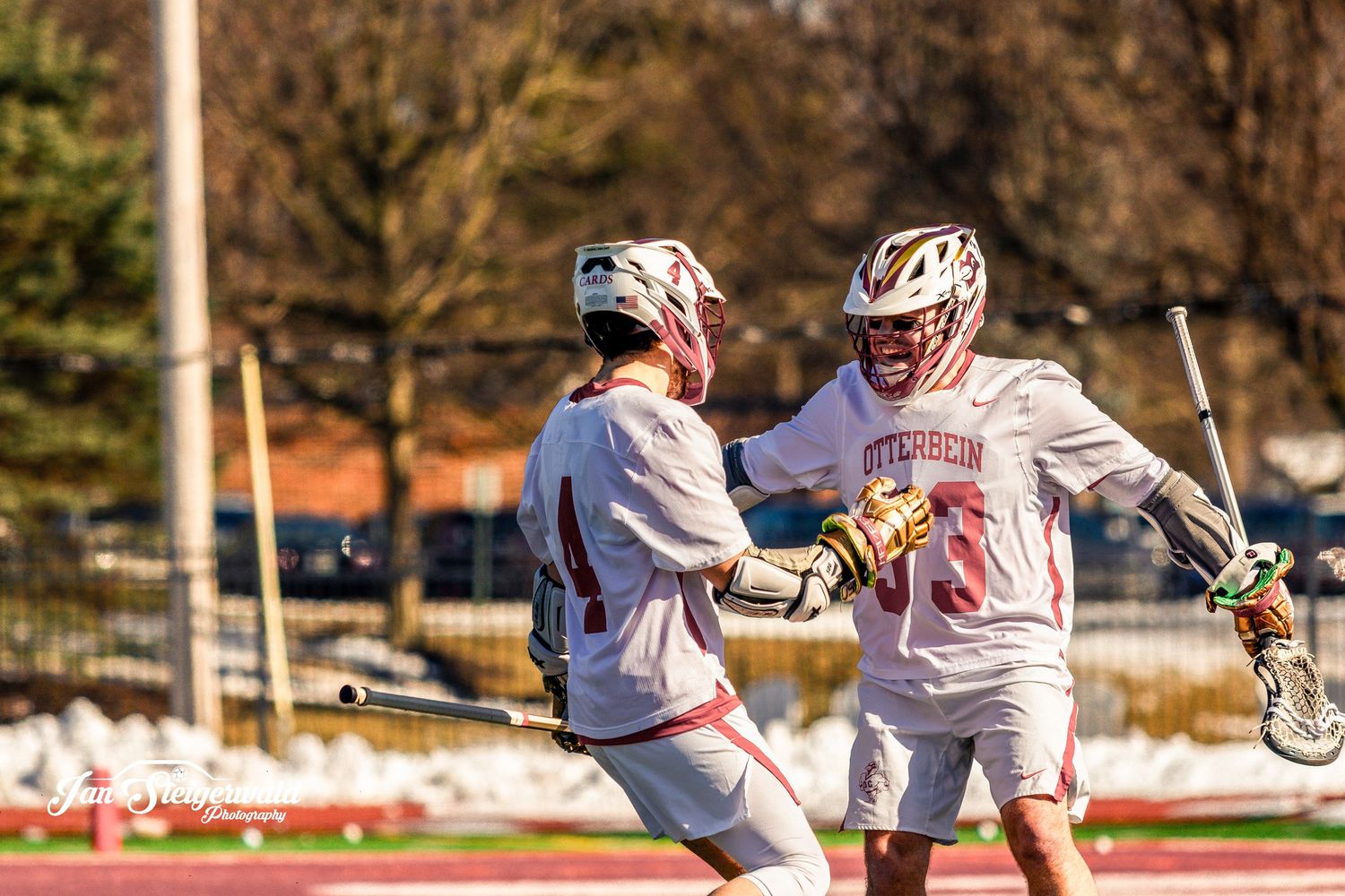 Two lacrosse players in white uniforms celebrate together on the field during a sunny afternoon game.