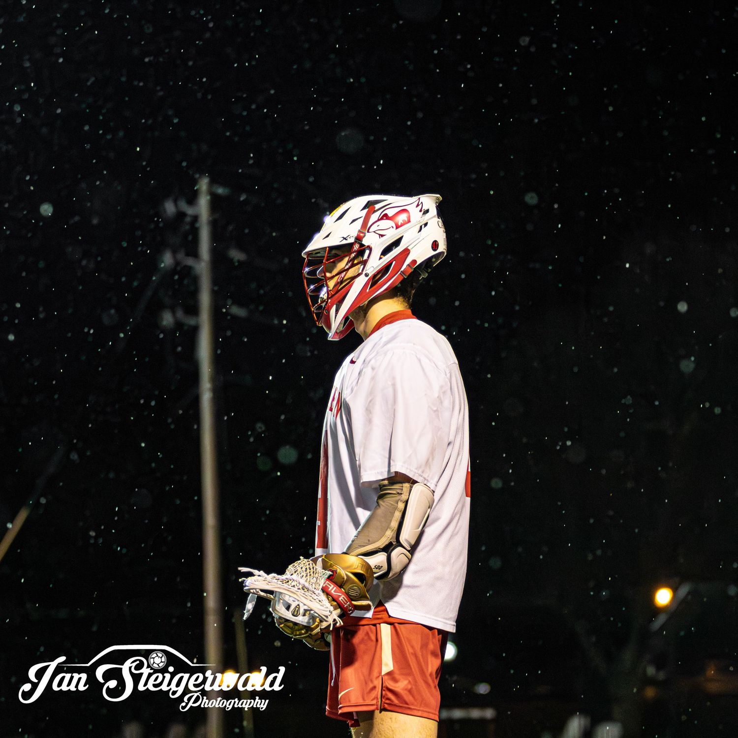 A lacrosse player wearing a white uniform and protective helmet stands against a dark starry night background.