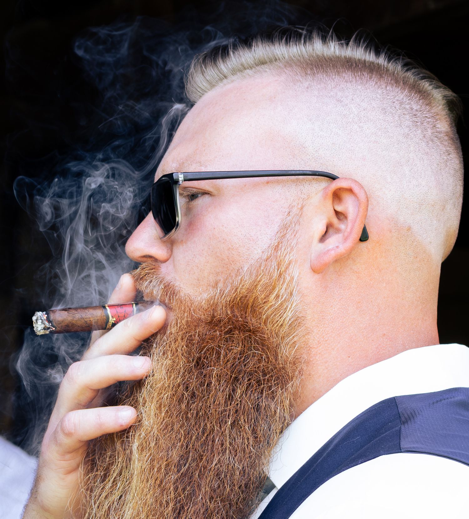 Side profile view of someone with a ginger beard smoking a cigar with rising smoke against a dark background.