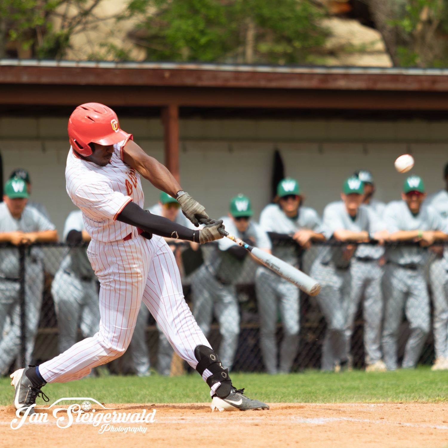 Baseball player in white pinstriped uniform and red helmet swings bat during game with team dugout visible in background.