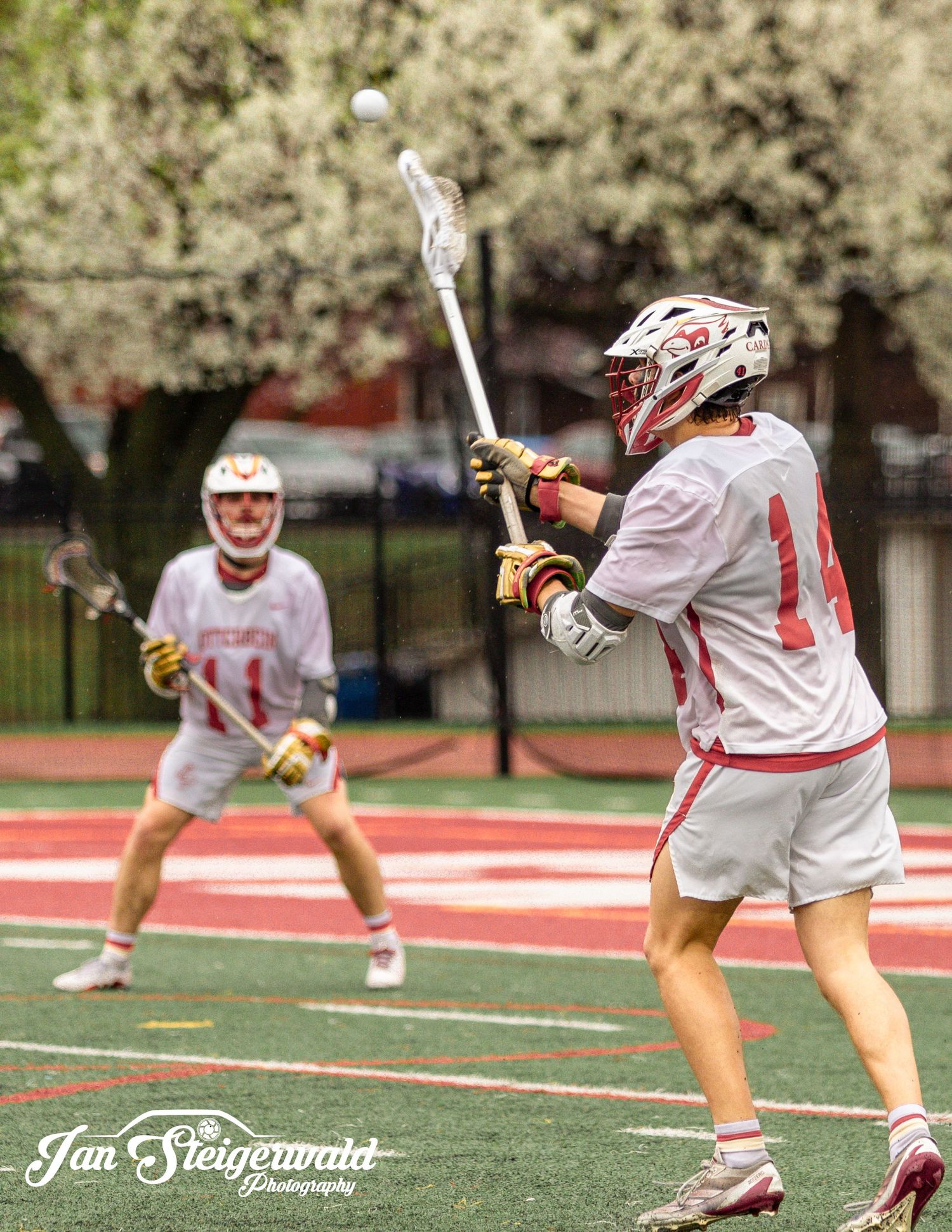 Lacrosse player in white jersey releases a shot on the field while teammate watches in the background.