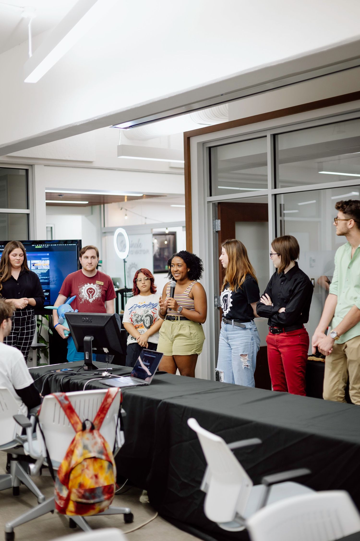Group of students gather around computers in a modern classroom with equipment and presentation displays visible.