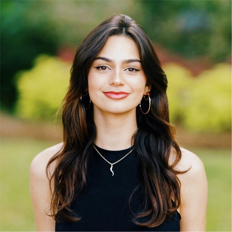 Woman in black sleeveless top with long dark hair and jewelry against blurred green background.