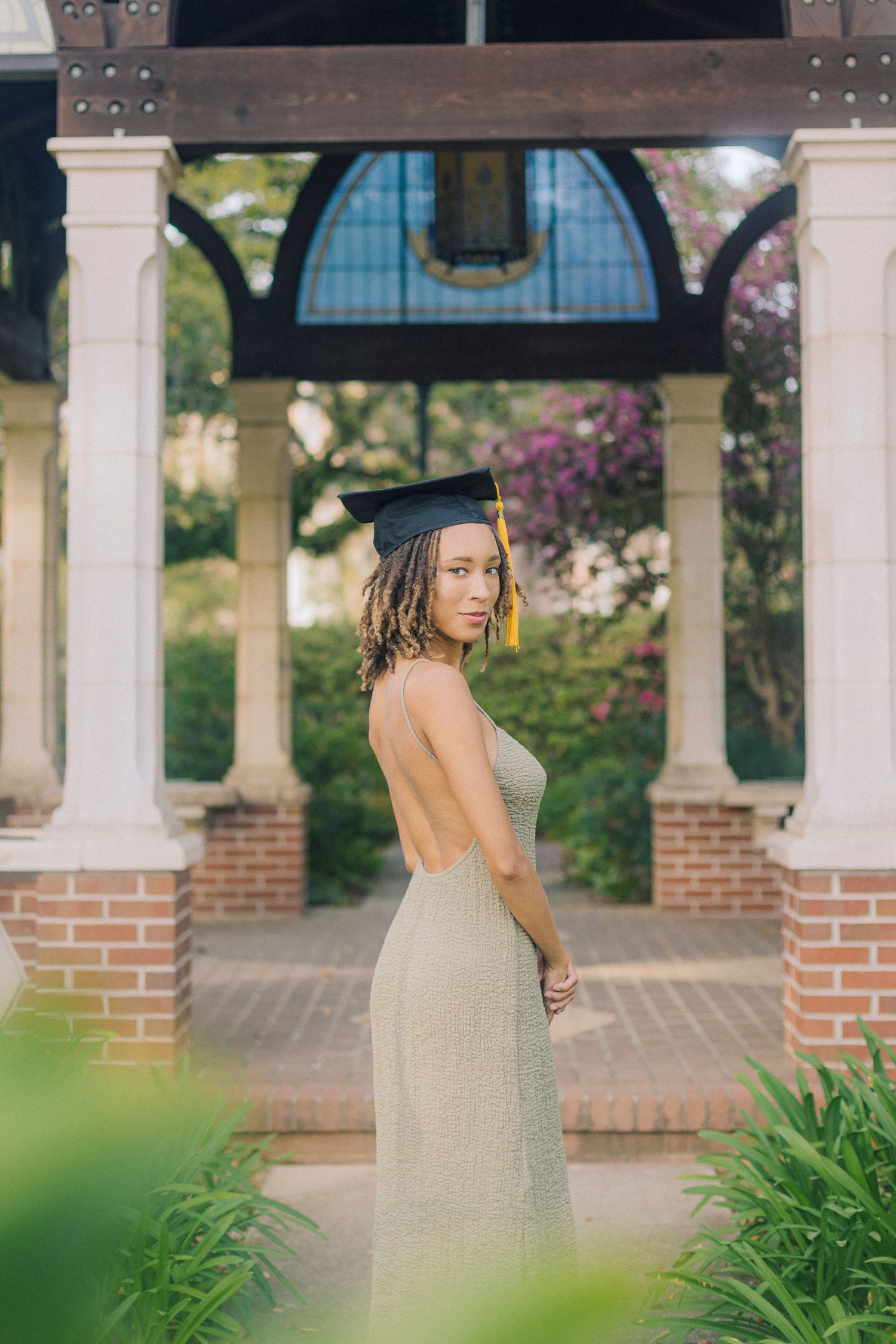 Graduate poses in sage green dress beside white columns and greenery in a classic outdoor setting at university campus.