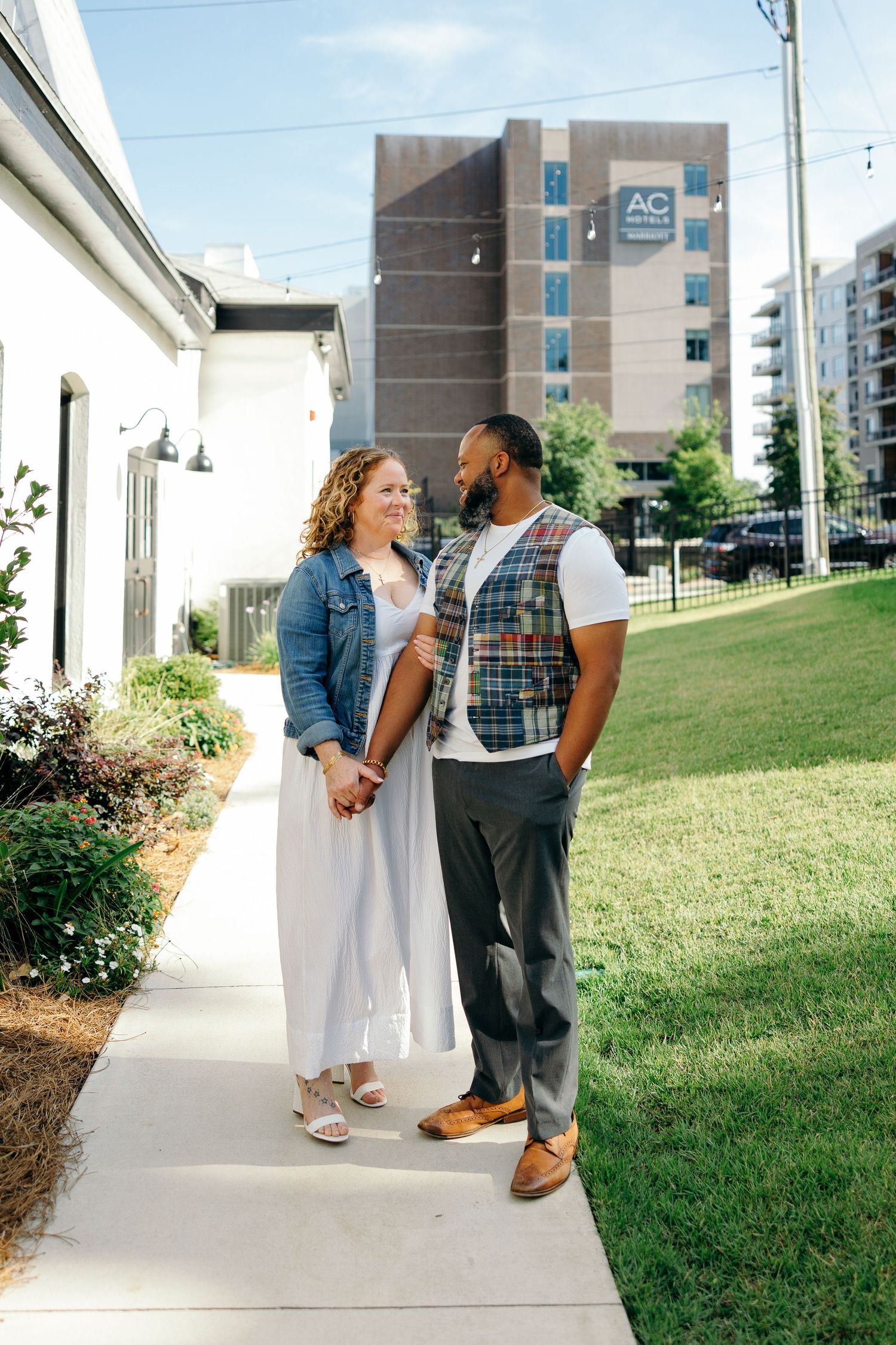 A couple stands together on a sidewalk with a modern apartment building in the background on a sunny day.