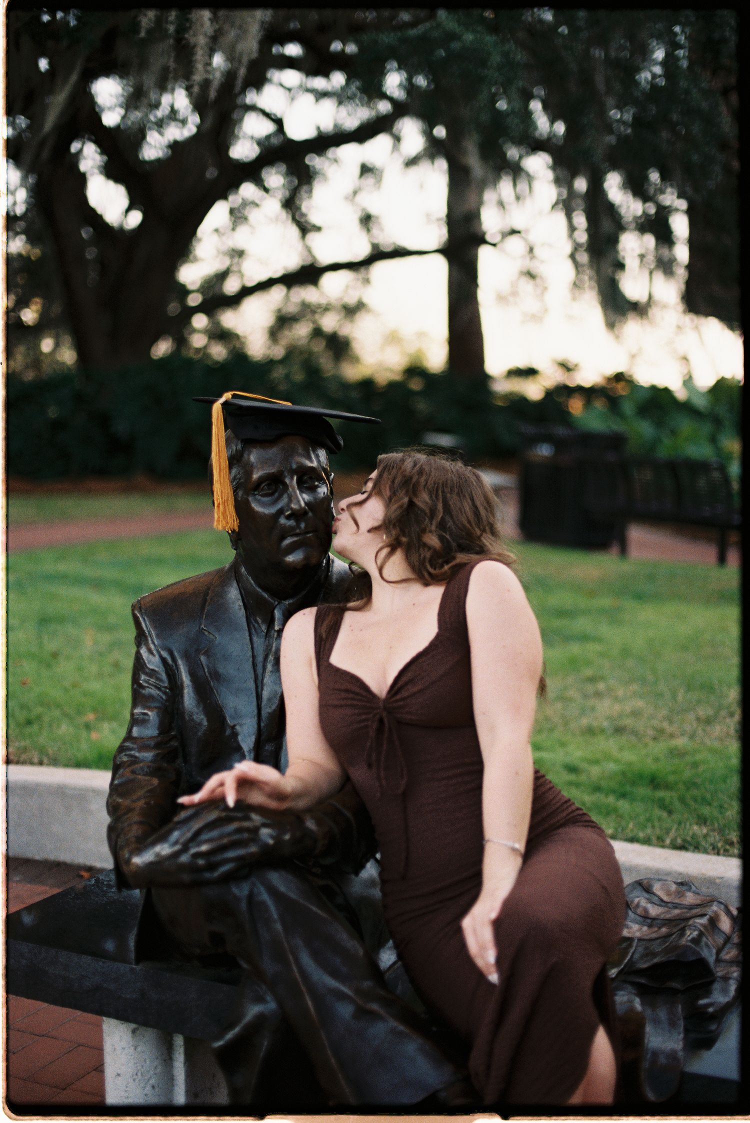 Woman in brown dress posing with a bronze graduation statue on a bench in a park at dusk.