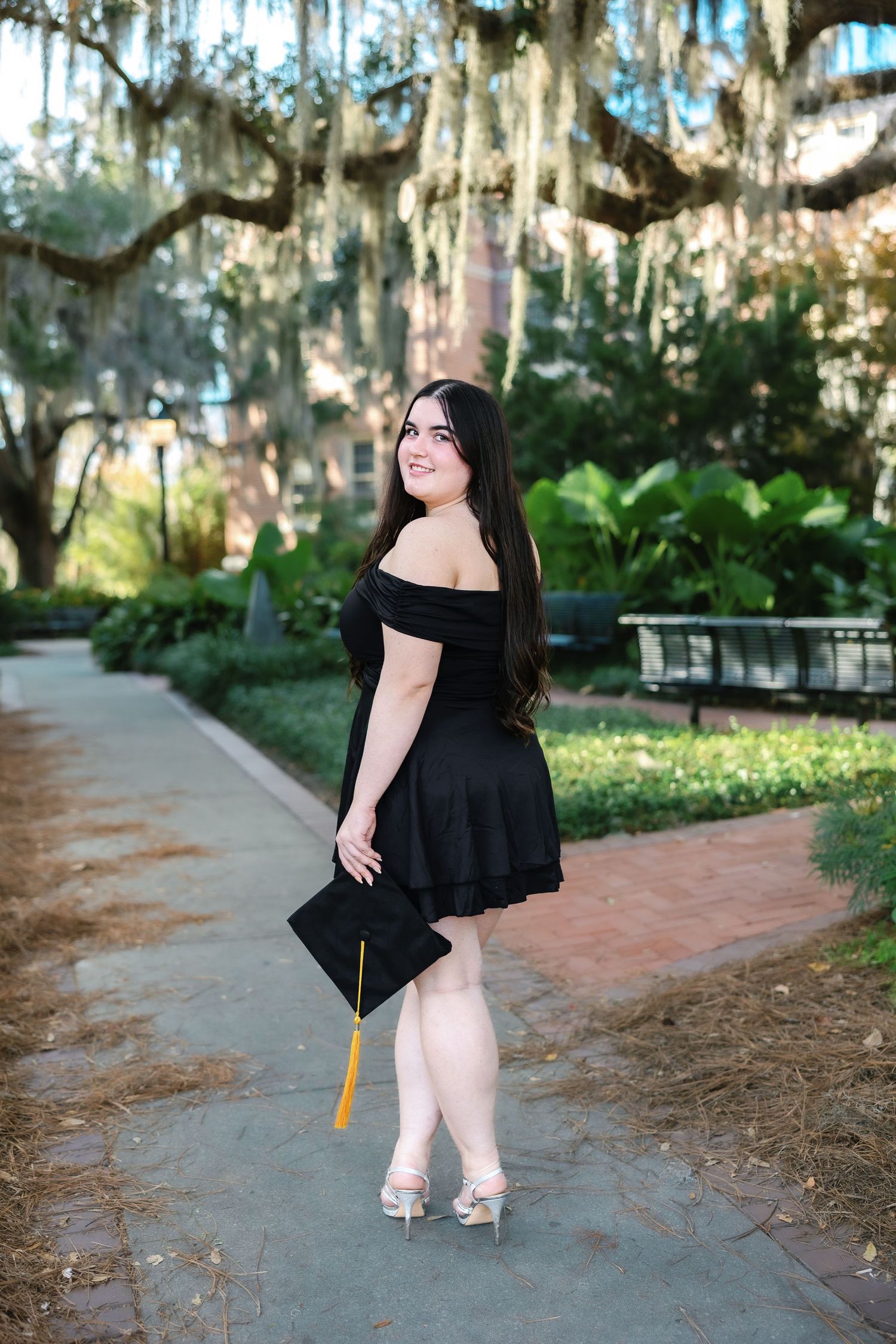 A person in a black dress poses gracefully among Spanish moss-draped trees on a brick pathway.