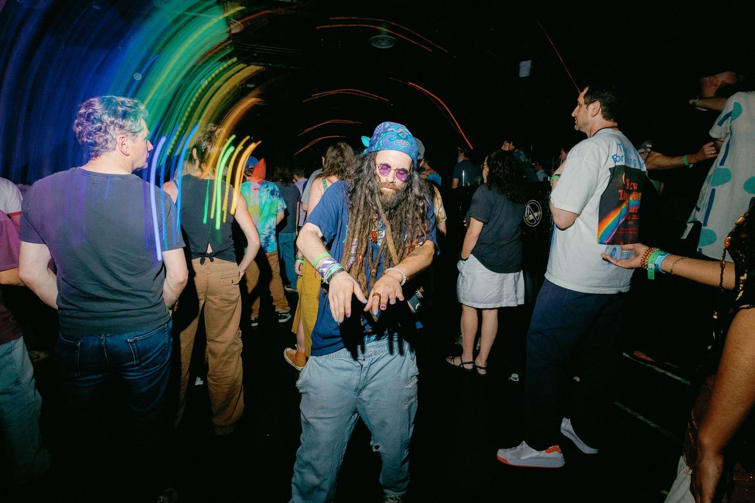 Man with dreadlocks dancing in colorful rainbow tunnel at a nightclub party event.