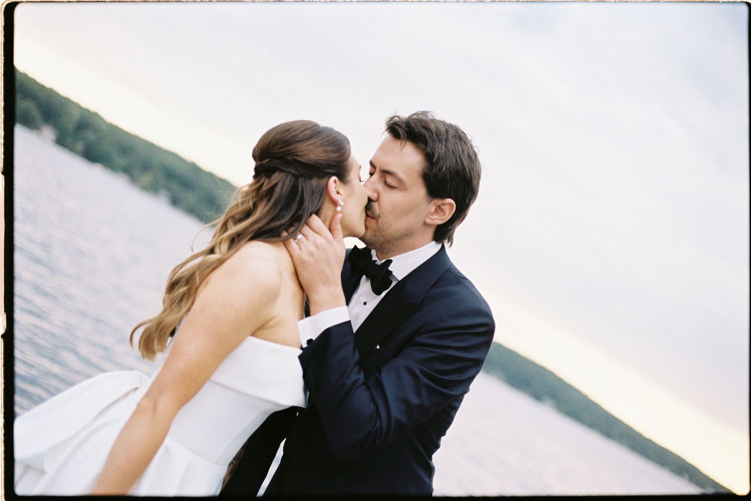 Bride and groom sharing a romantic kiss by a lakeside at sunset, with the groom in a navy tuxedo.