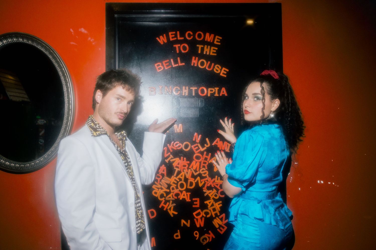 Two people pose by a neon sign reading Welcome to the Bell House Synchtopia on an orange wall.