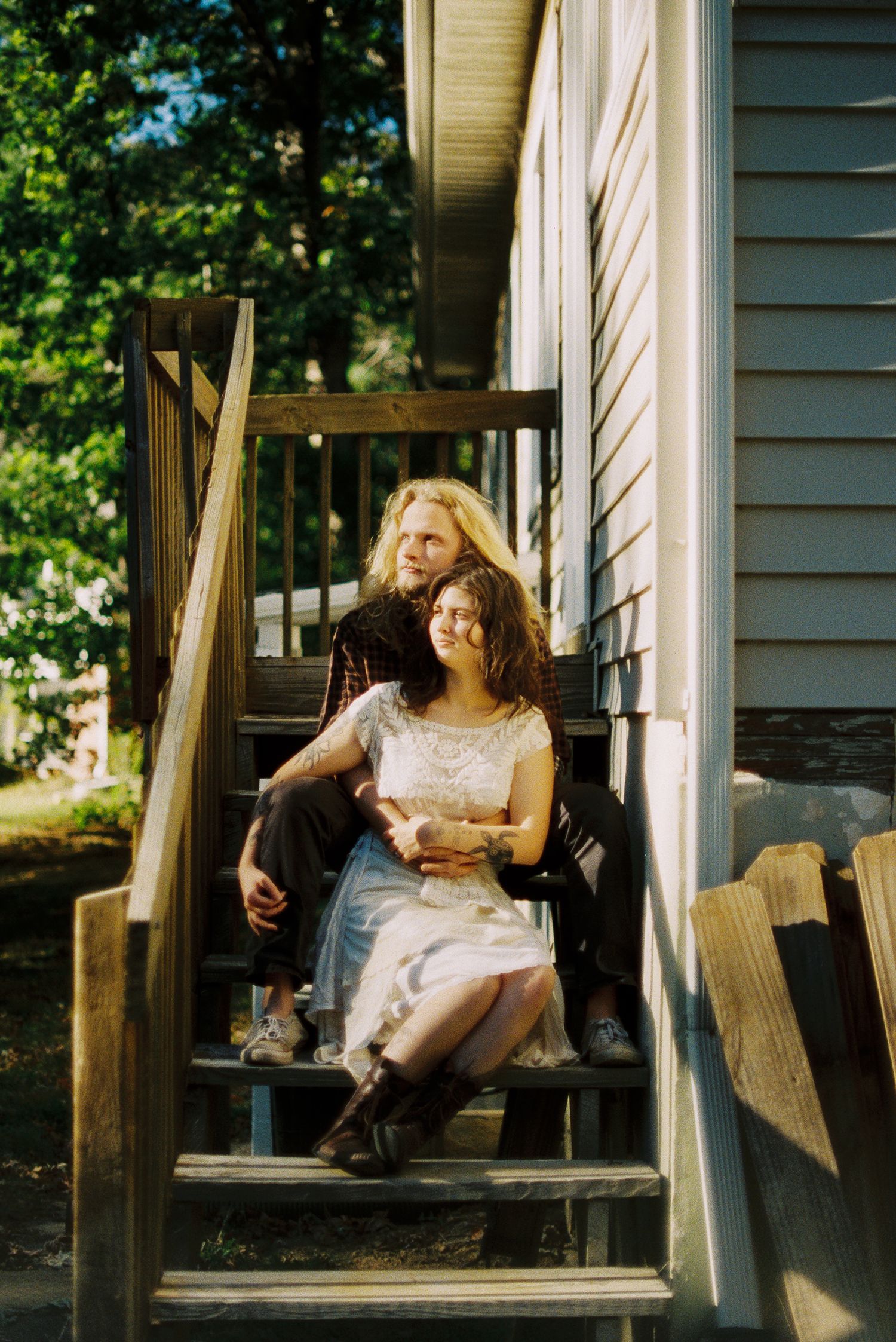 Two women sit close together on wooden porch steps beside a house with gray siding, surrounded by green trees in warm sunlight.