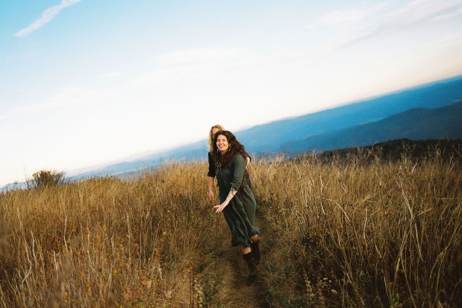 Two women walk along a narrow path through golden tall grass on a mountain ridge with blue hazy peaks and a soft sky behind them.