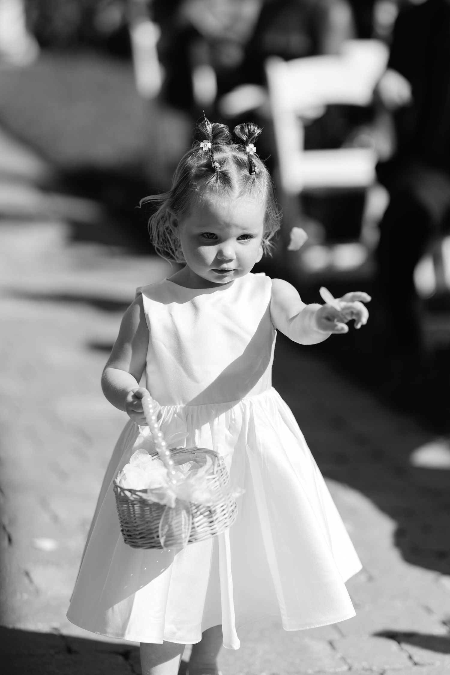 Black and white photo of a young flower girl in white dress carrying a basket, tossing petals at a wedding.