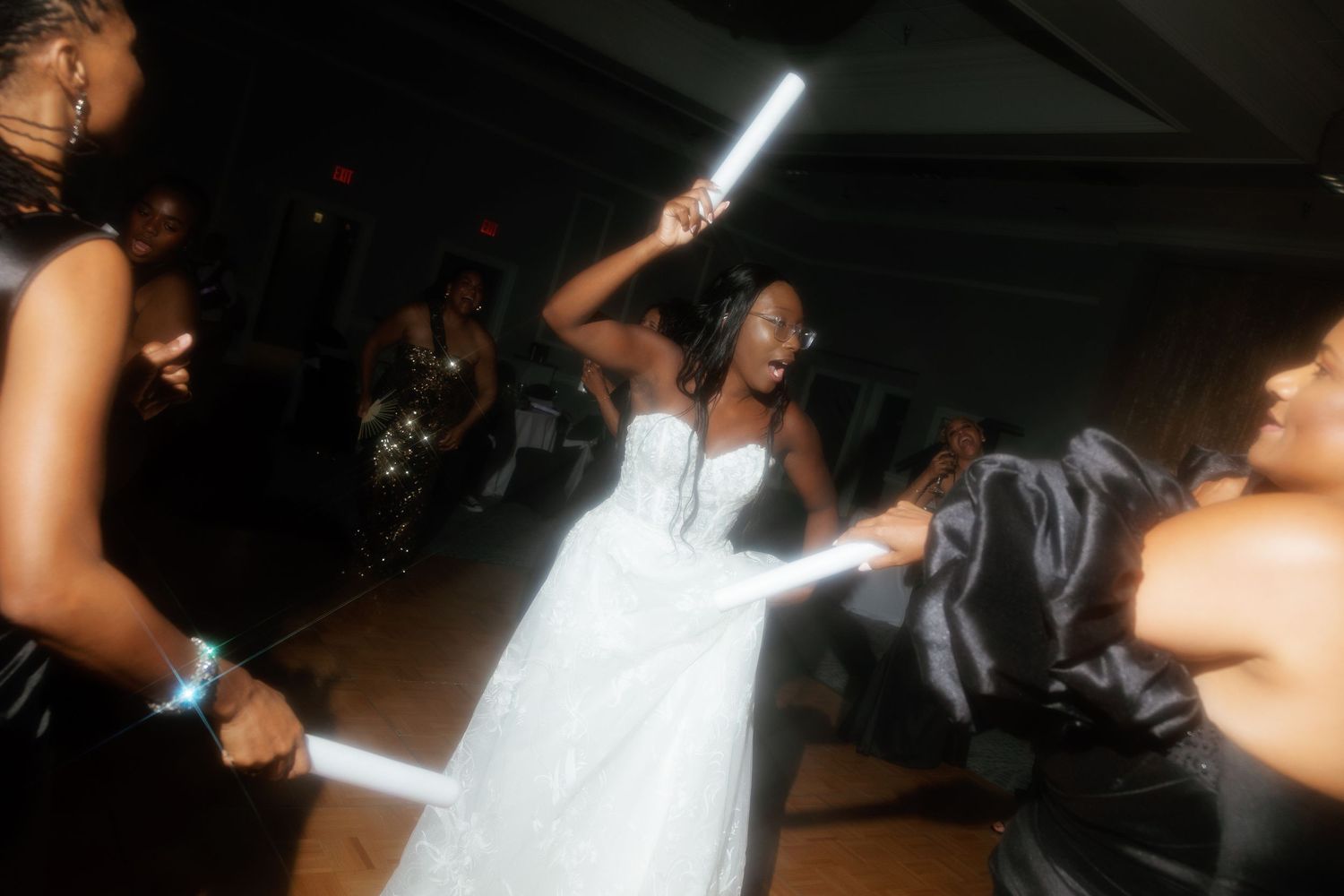 Bride in white gown dancing joyfully with guests at a wedding reception, holding a glow stick.