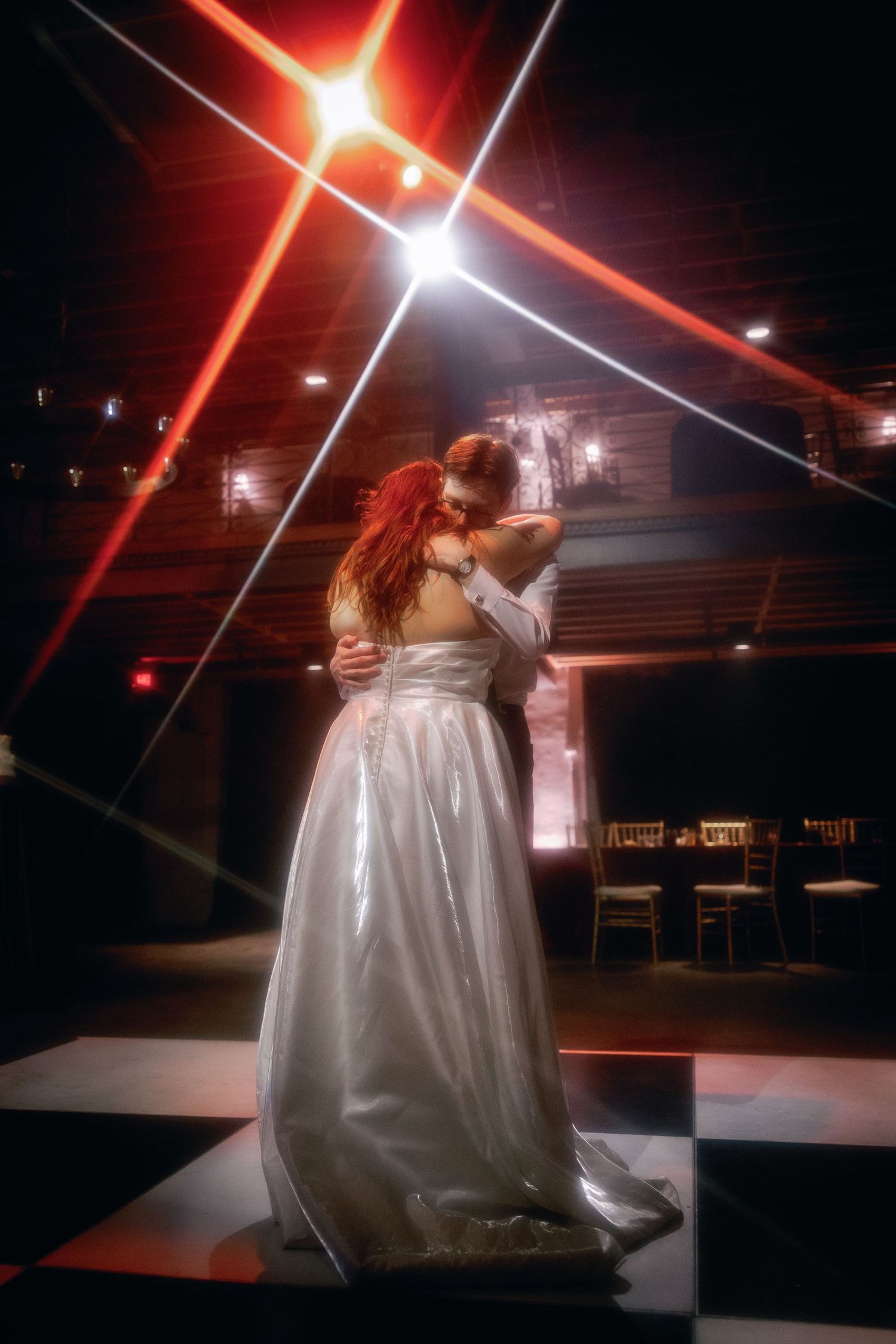 Bride and groom share first dance on checkered floor under dramatic red laser lights at wedding reception.
