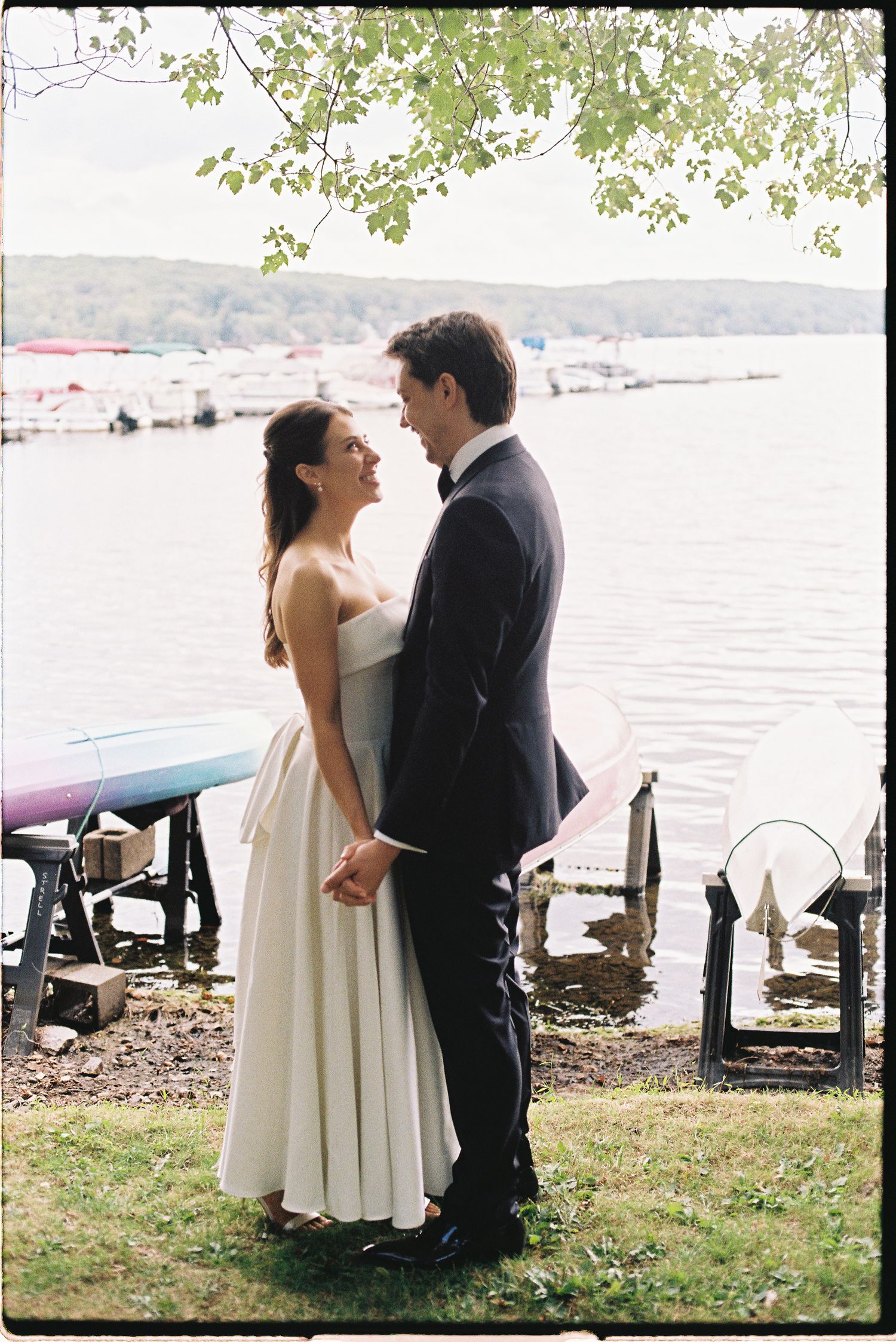 Bride and groom holding hands by a lakeside dock, smiling at each other in wedding attire on a cloudy day.