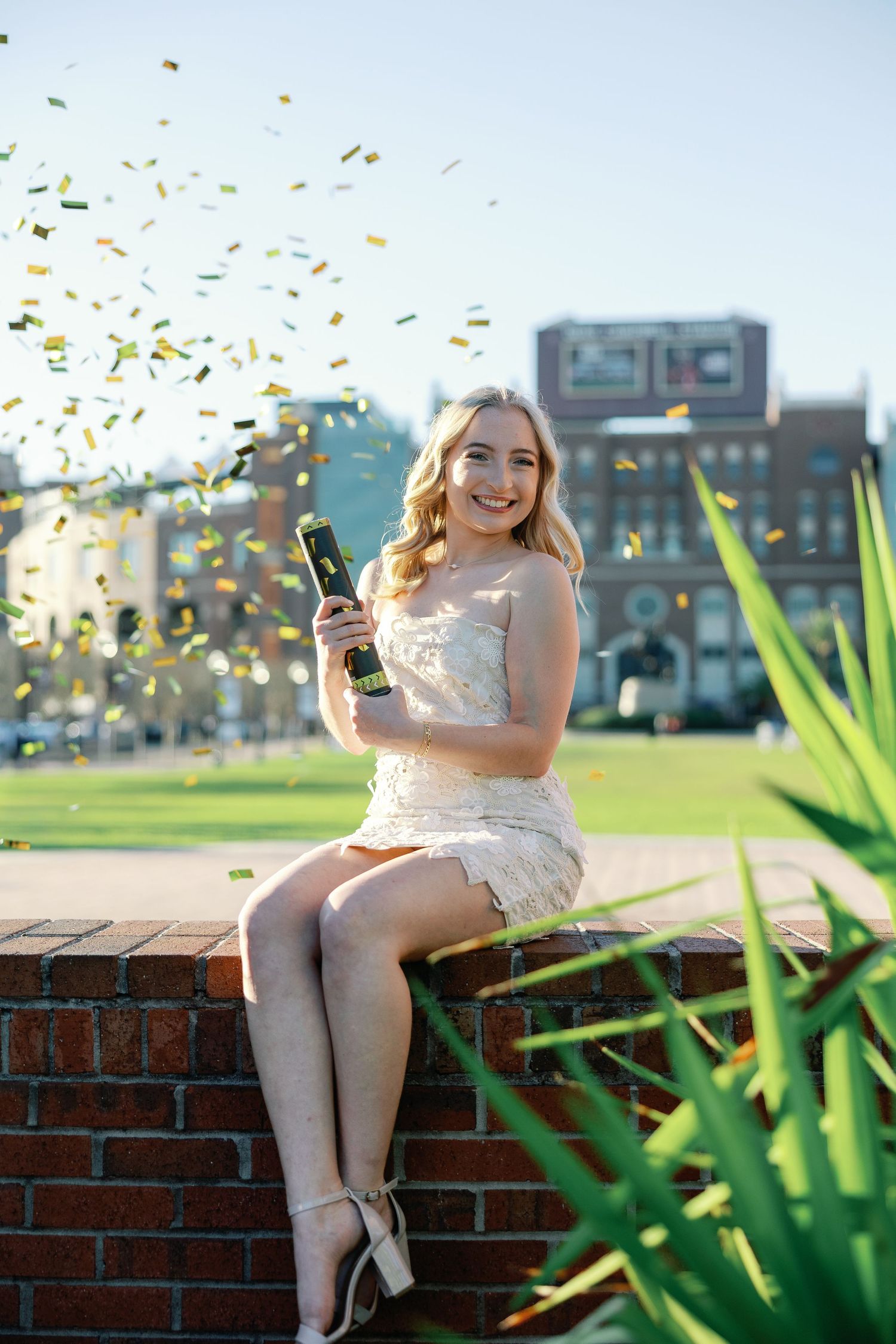 A person in a white lace dress sits on a brick wall while confetti falls during a celebratory outdoor moment.