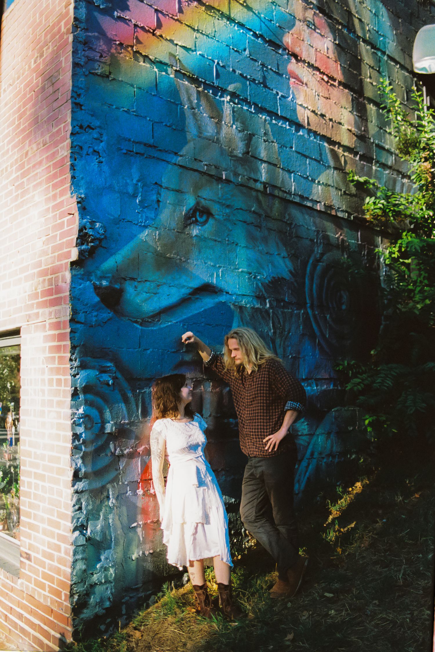 A couple in casual clothing leans together against a brick wall featuring a vibrant blue wolf mural, surrounded by green plants.