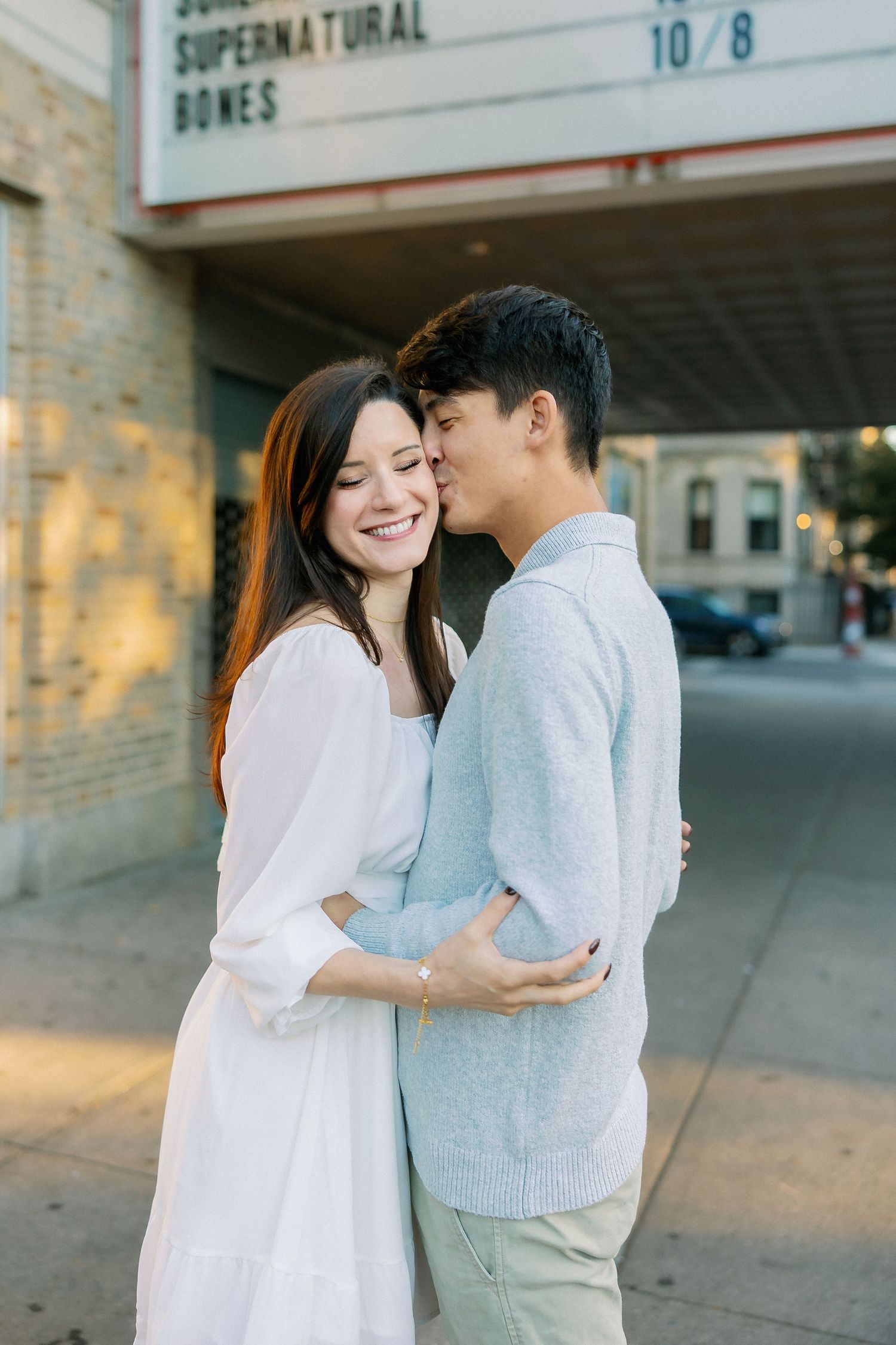 A couple in white and light blue outfits share tender moments together against a brick wall during a photo session.