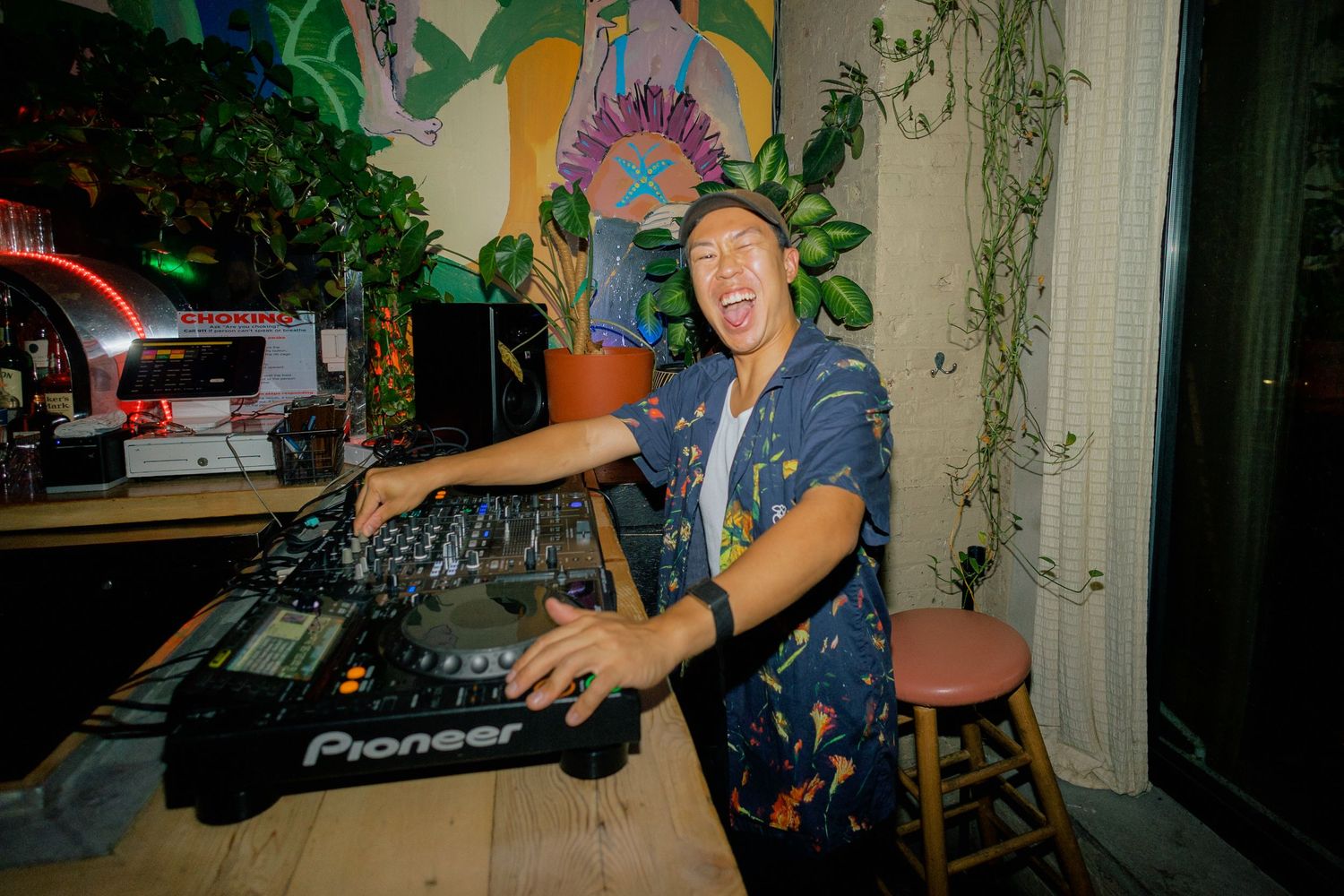 Smiling DJ mixing music on Pioneer turntables at a tropical-themed indoor venue with lush green plants.