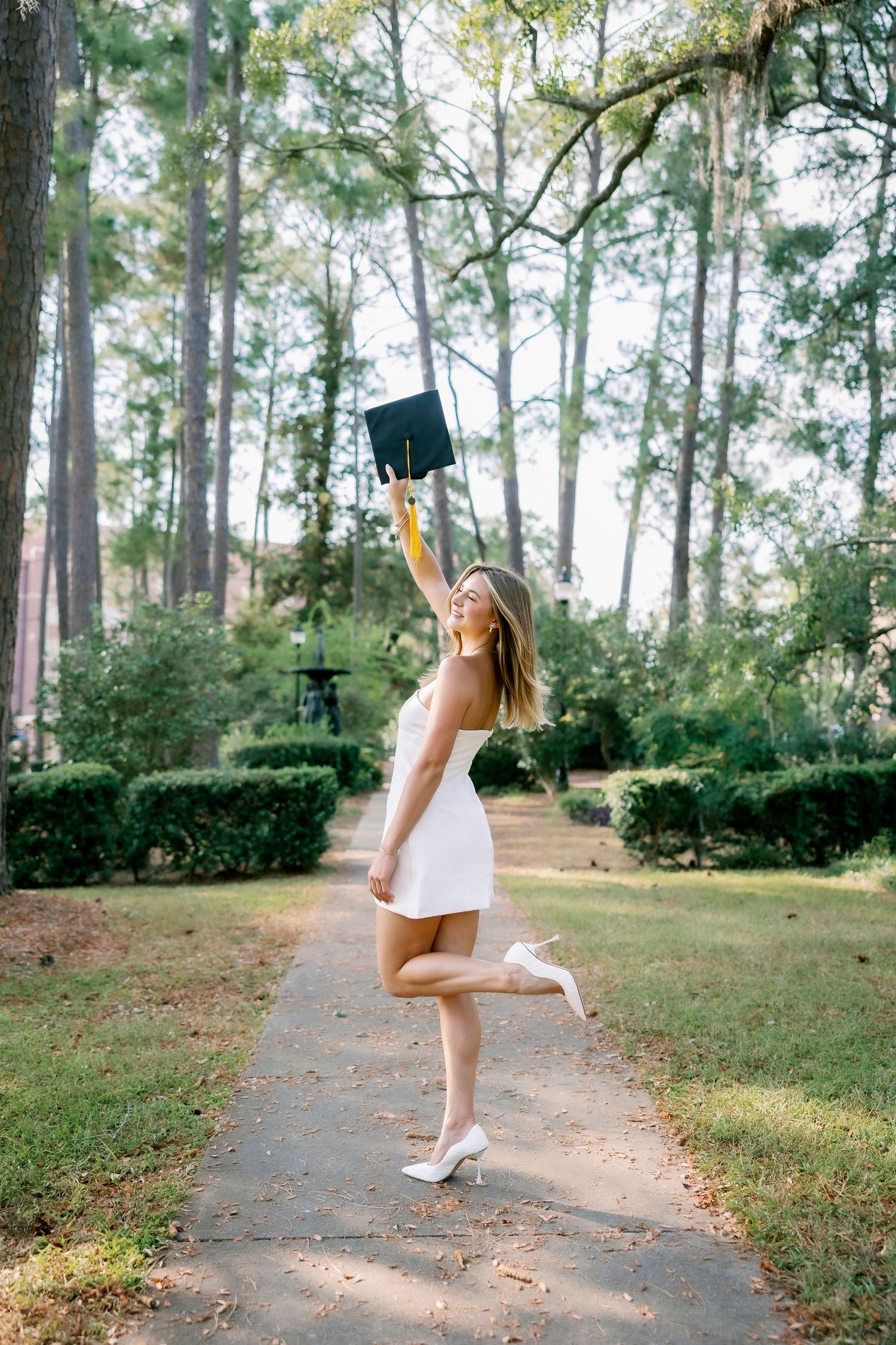 A person in a white dress dances and spins joyfully along a tree-lined path in a sunlit forest setting.