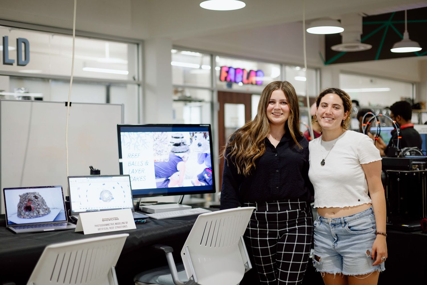 Two people standing in a modern office space with computer monitors and workstations visible in the background.