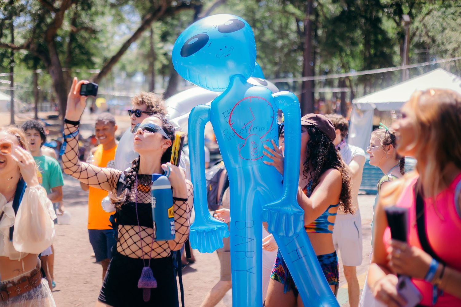 A large blue inflatable alien figure stands among festival attendees at an outdoor summer event.