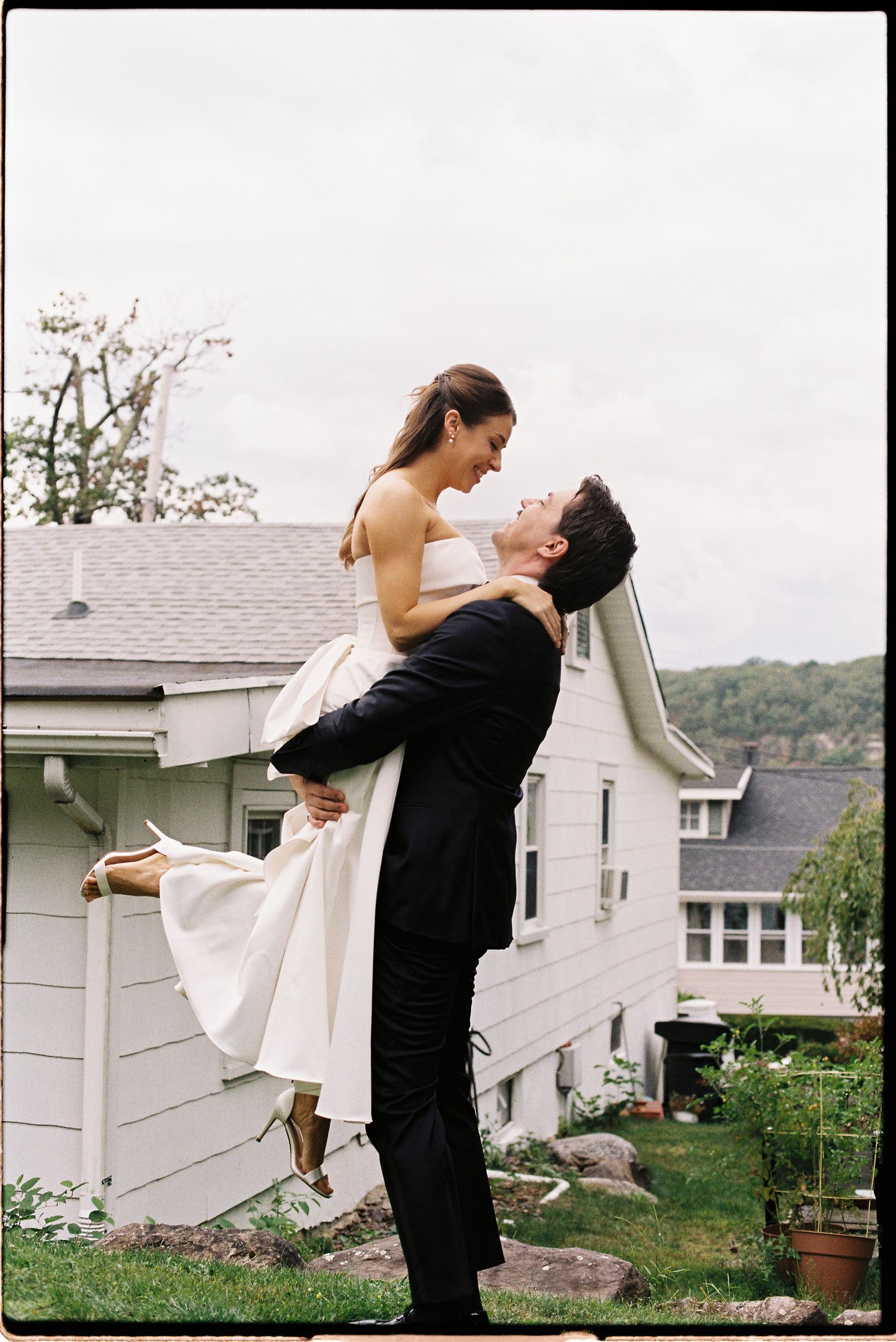 Groom lifts bride in white dress outdoors near house on wedding day.