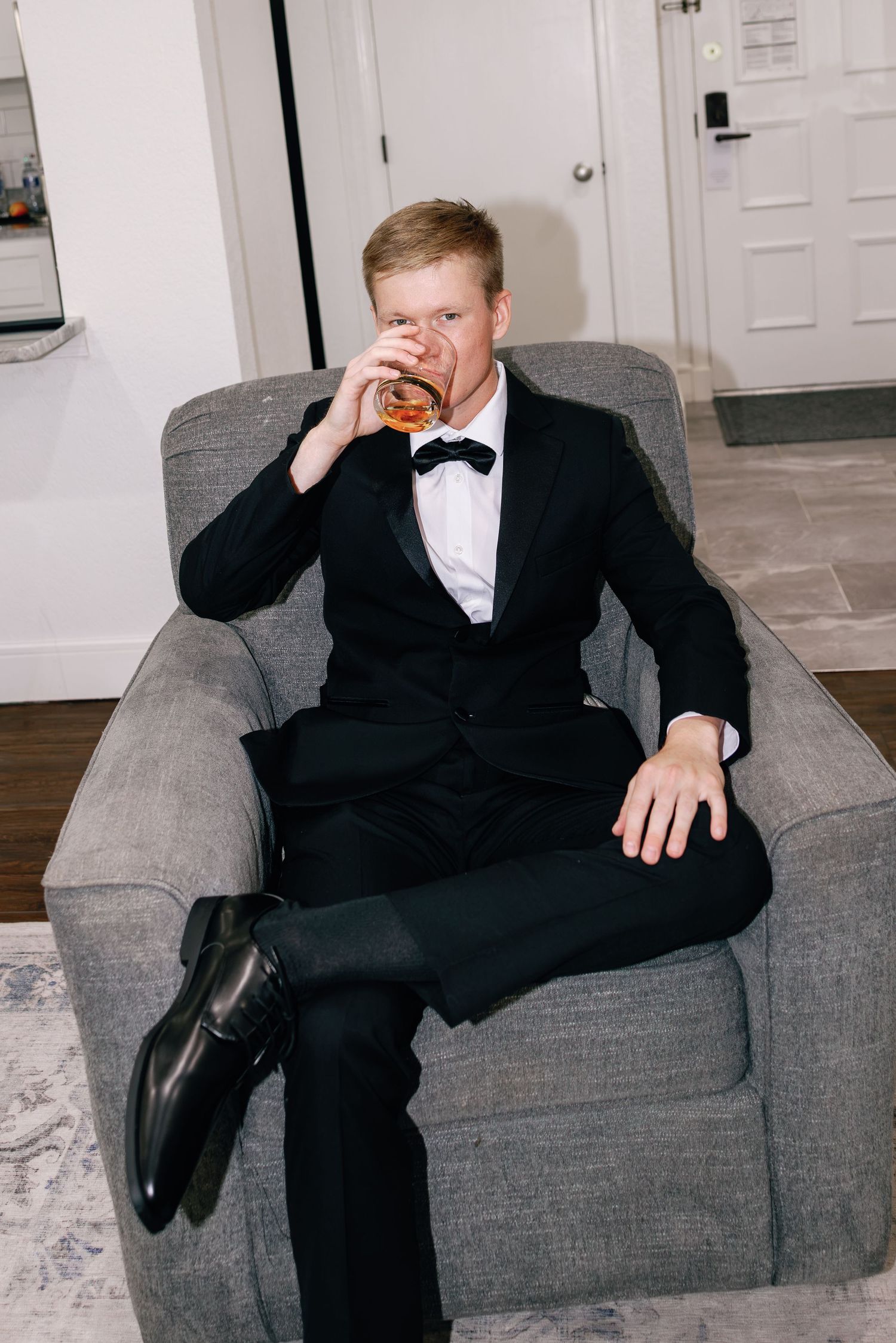 Groom in black tuxedo with bow tie sipping whiskey while seated in gray armchair before wedding ceremony.