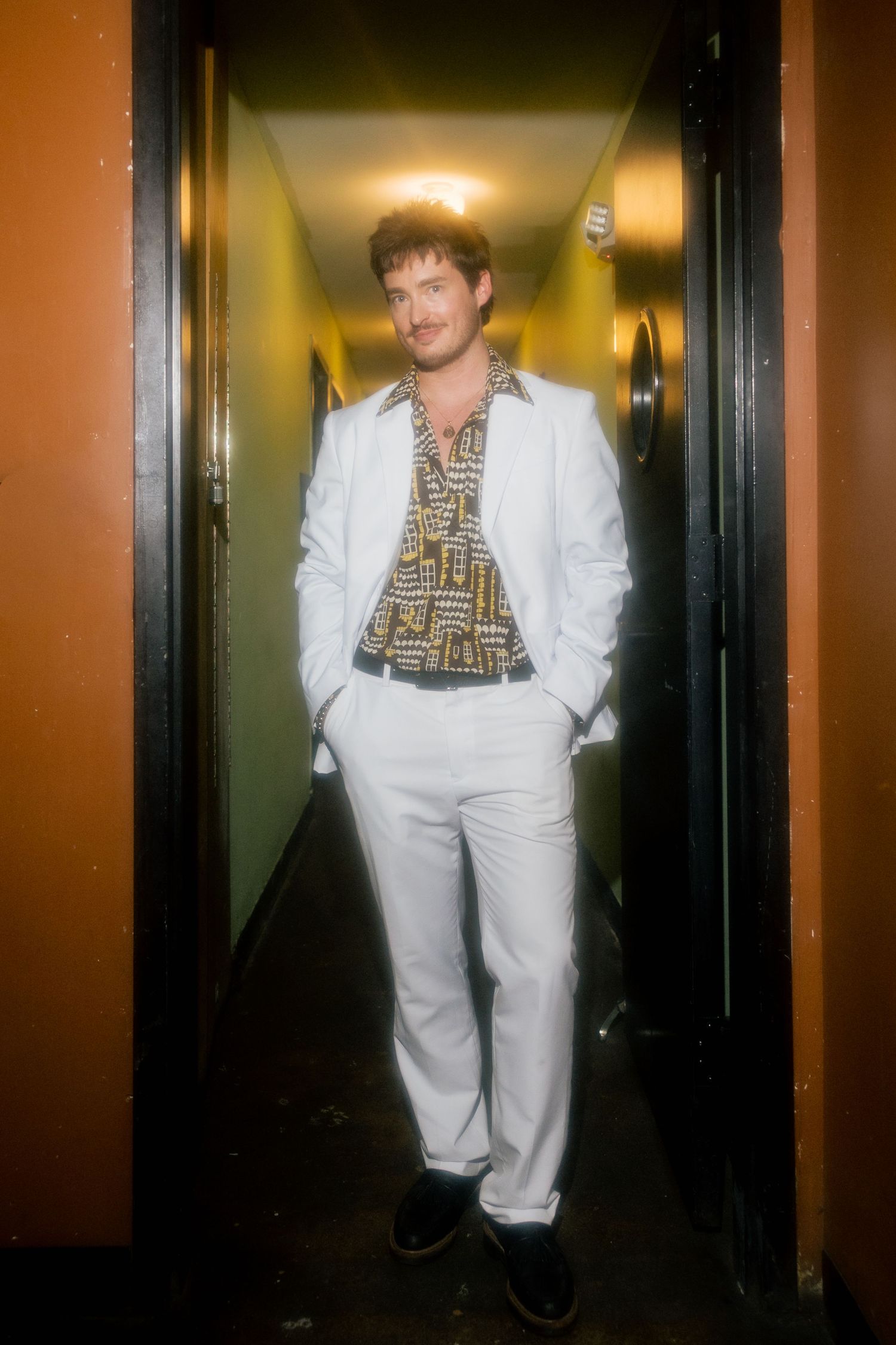 Man in white suit and patterned shirt standing in a doorway of an orange hallway.