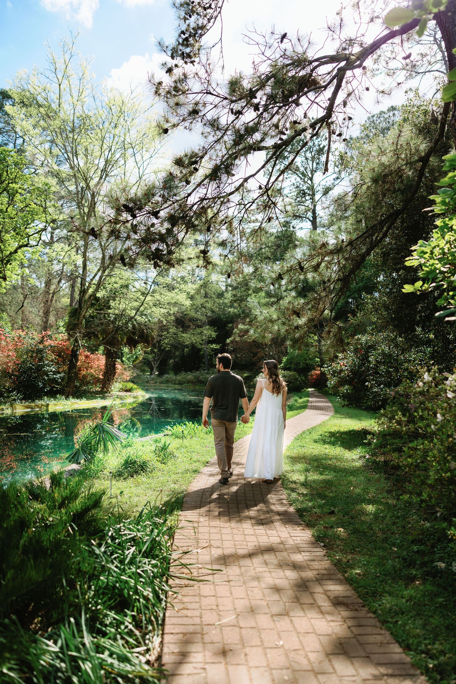 A couple walks along a winding brick path through a lush garden with blooming trees and a tranquil pond.