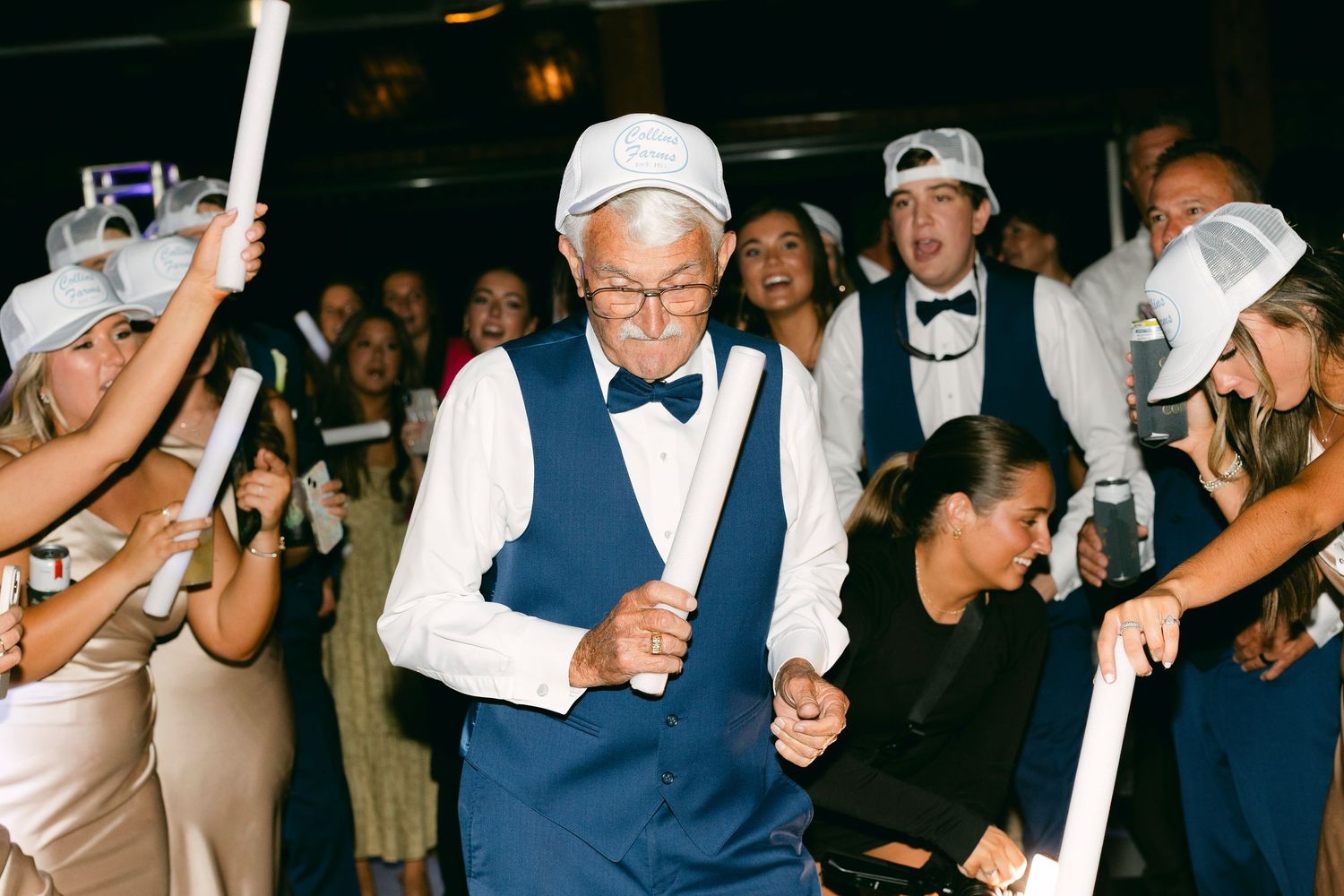 Wedding guests dance and celebrate on a dark dance floor with glowing light sticks during an evening reception.