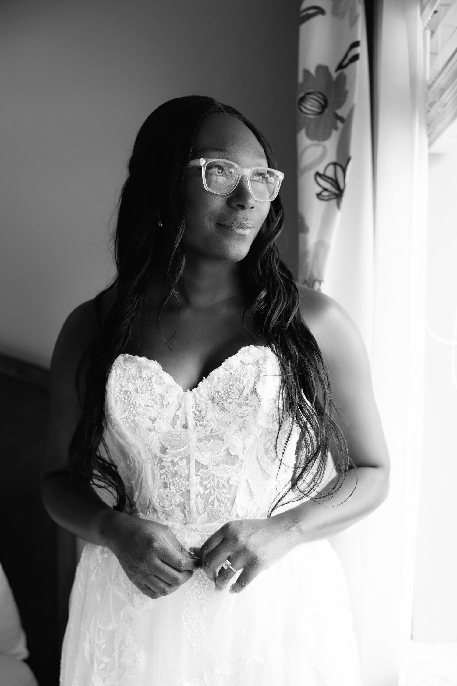 Black and white portrait of bride in strapless lace gown wearing white glasses by window.