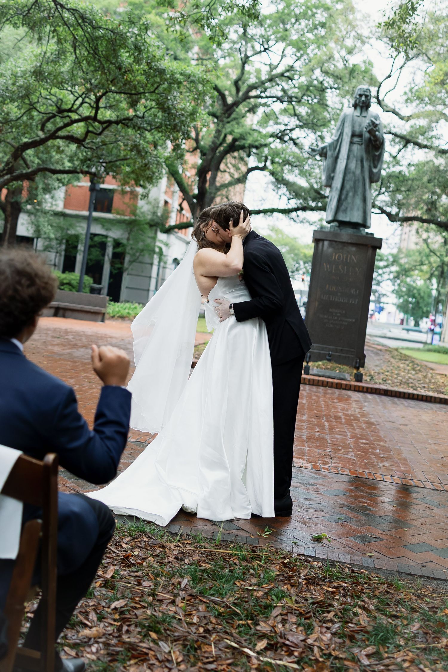 Bride and groom share first kiss near bronze statue in rainy outdoor park while wedding guest watches.