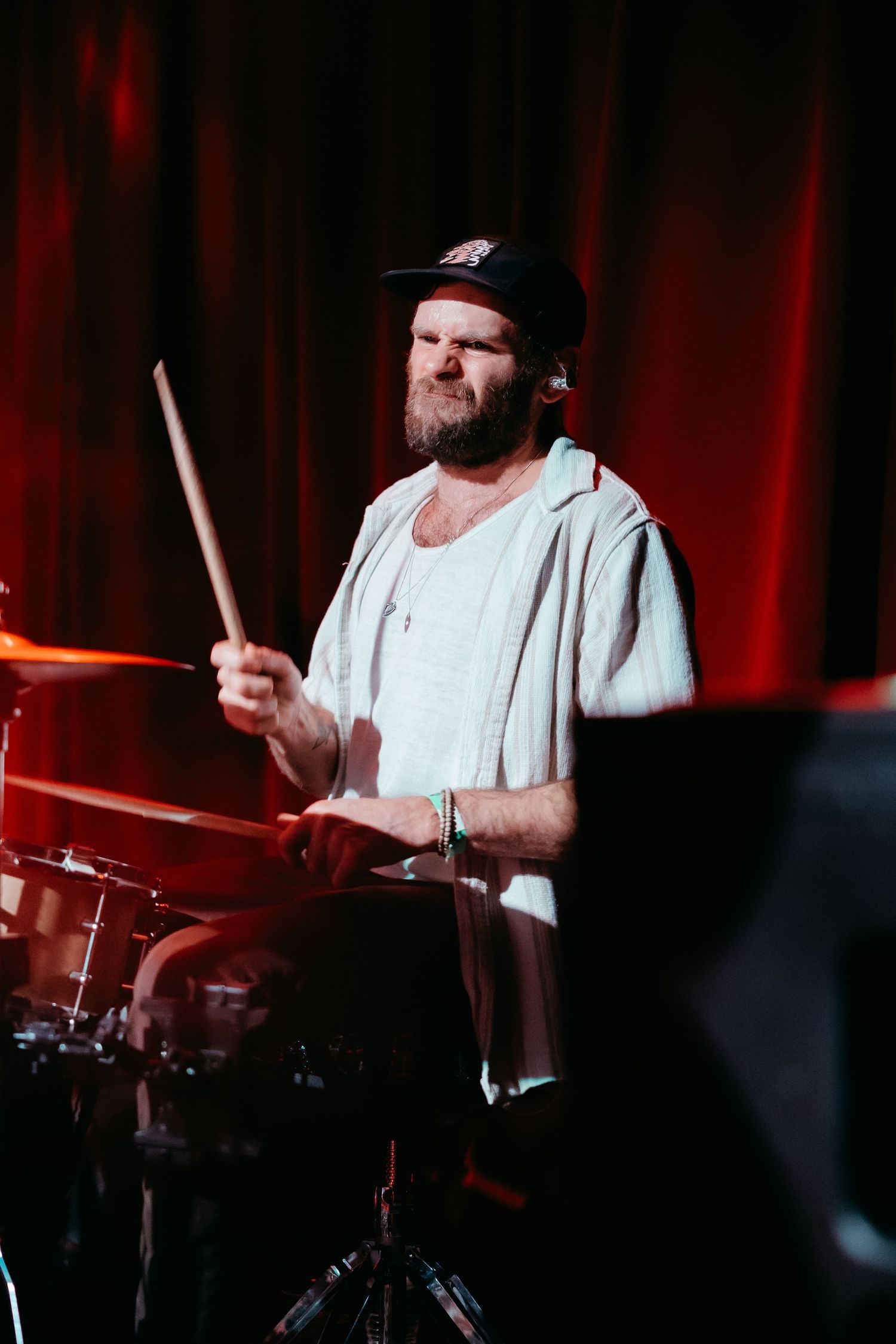 Drummer in a white shirt performs energetically on stage with red lighting in the background.