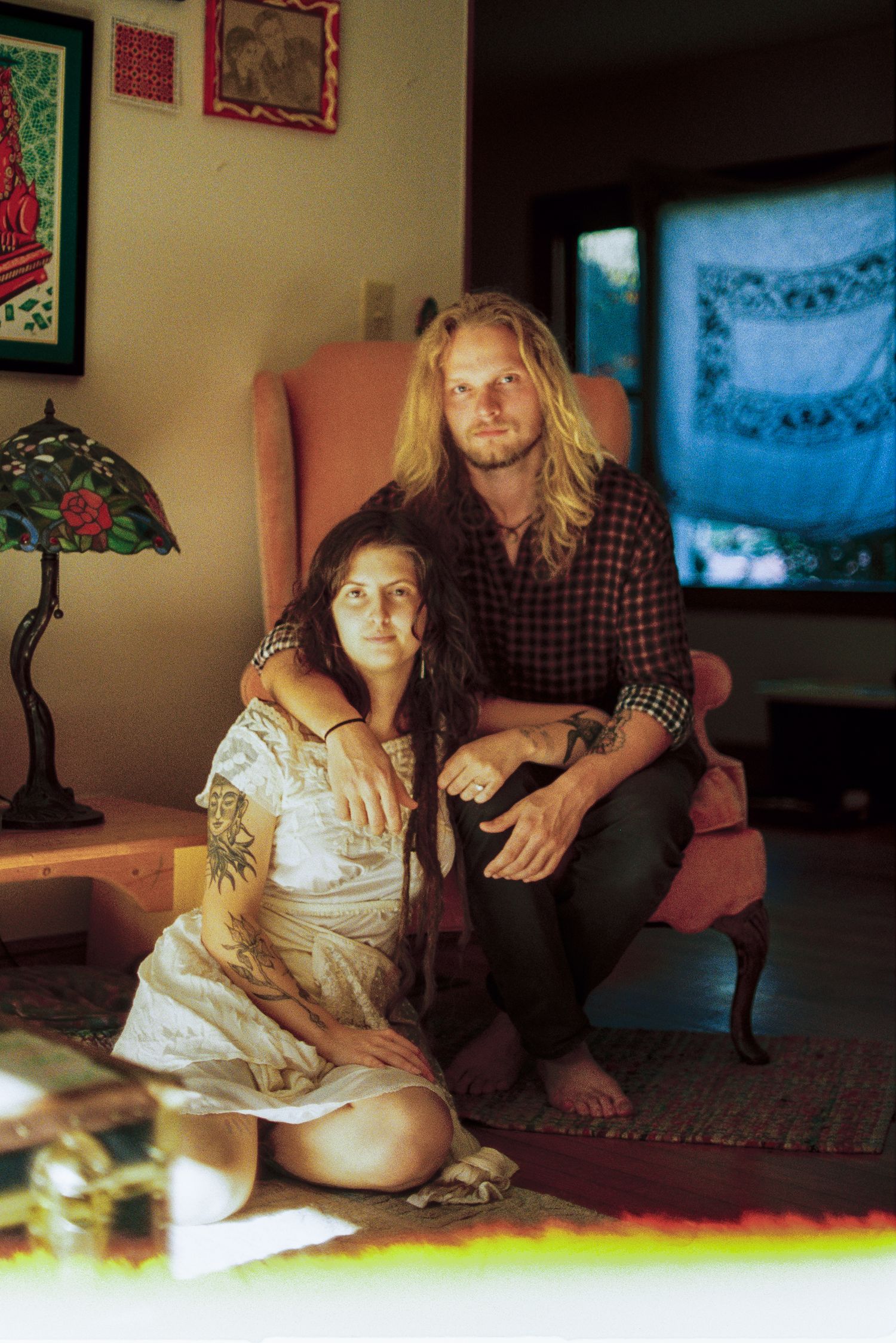 A bearded man with long blond hair sits in a pink armchair, embracing a tattooed woman seated on the rug in a cozy, art-filled room.