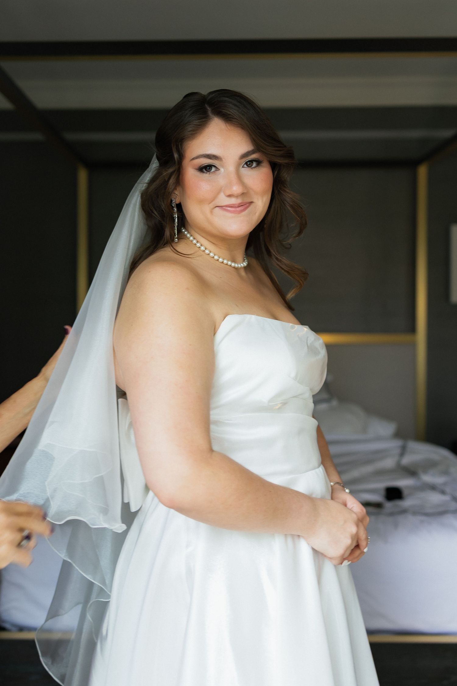 Smiling bride in strapless white gown and veil with pearl necklace getting ready in elegant room.