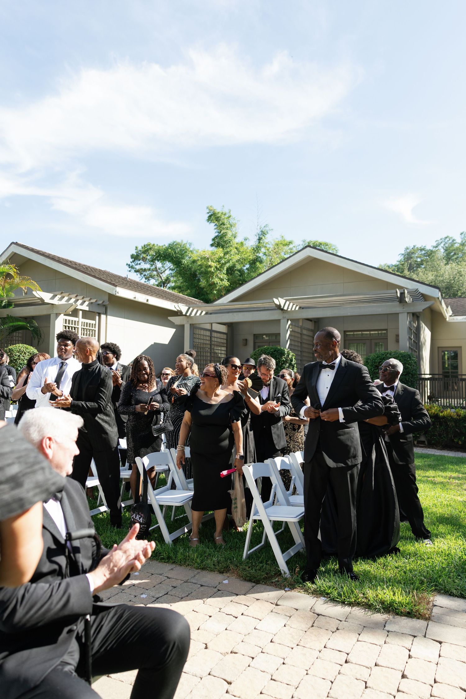Guests in black formal attire gather outdoors for an elegant wedding ceremony on a sunny day.