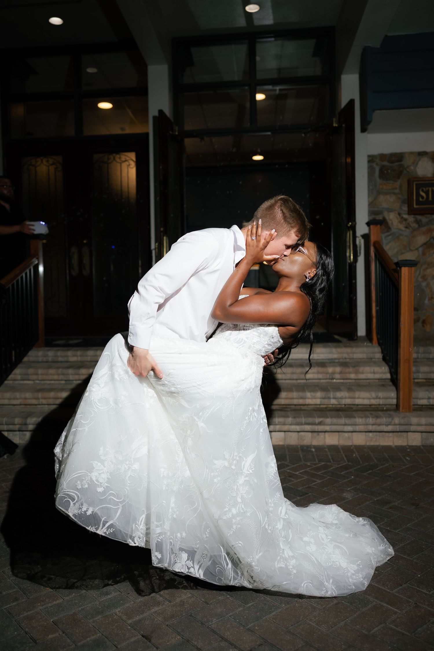 A groom dips his bride in a romantic kiss outside a venue, her white lace wedding gown flowing gracefully.