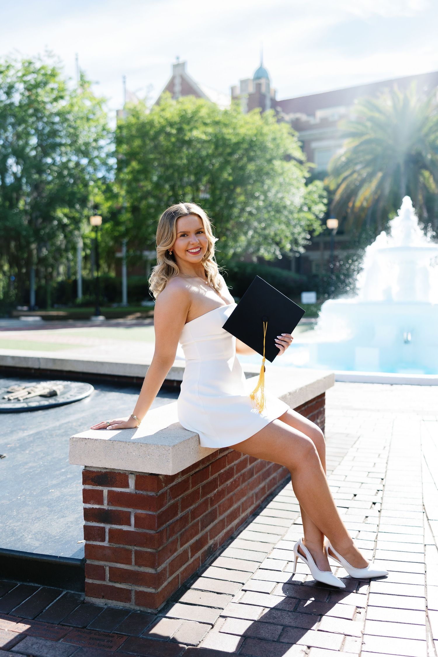 Young woman in white dress holding graduation cap sits on brick wall near fountain on sunny campus day.