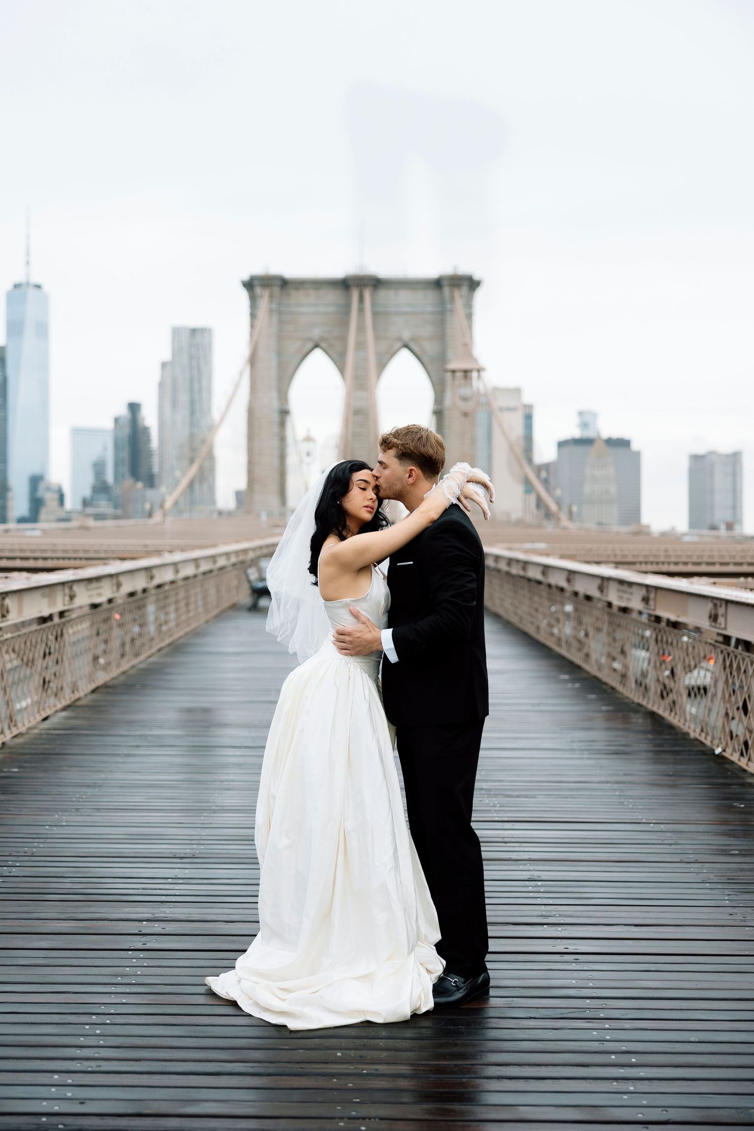 A couple in wedding attire shares romantic moments on the iconic Brooklyn Bridge with the NYC skyline in the background.