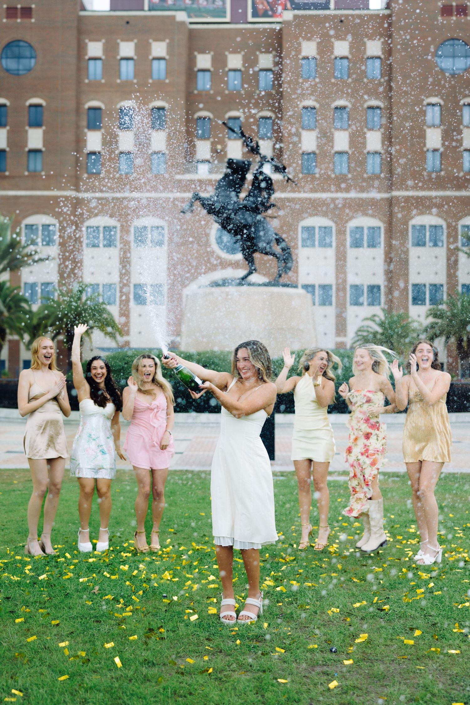Group of women celebrating outdoors with champagne spray near a horse statue on a university campus.