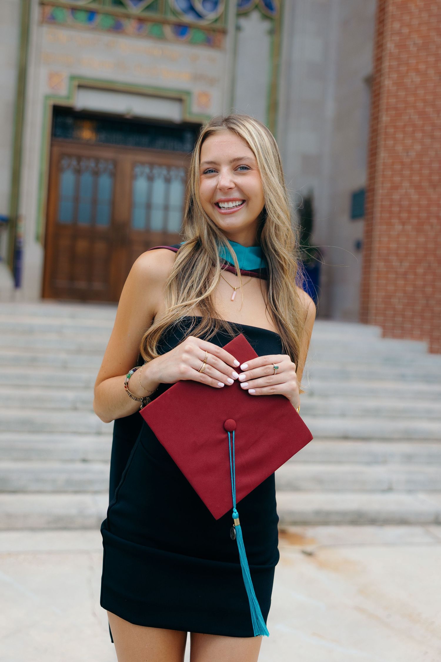 A college graduate poses with a red graduation cap in front of a brick building on campus.